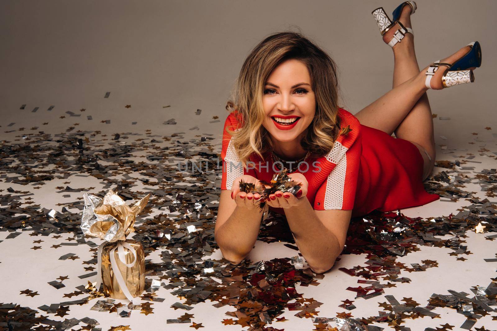 A girl in a red dress is lying on the floor with a bunch of gifts on a gray background in confetti by Lobachad