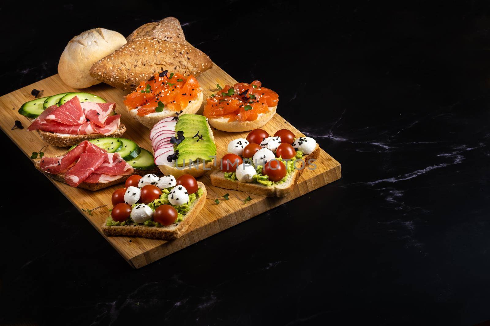 An assortment of sandwiches with fish, cheese, meat and vegetables lay on the board and a bun by Lobachad