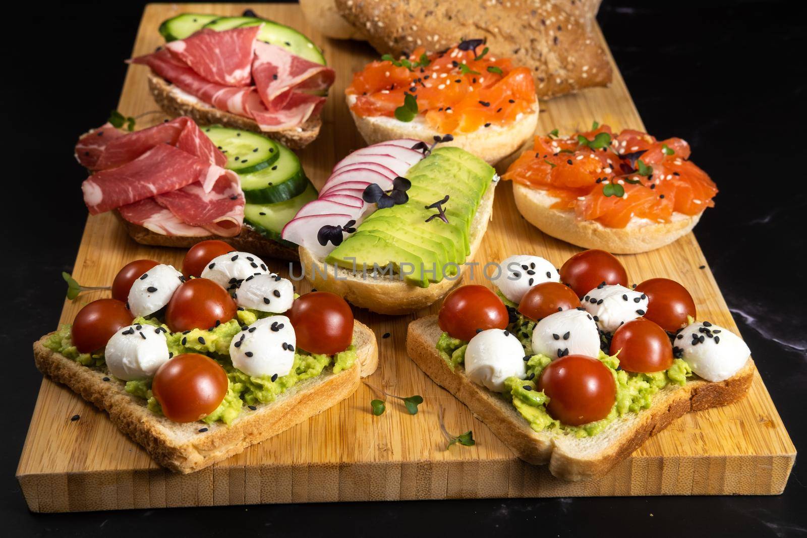 An assortment of sandwiches with fish, cheese, meat and vegetables lay on the board and a bun by Lobachad