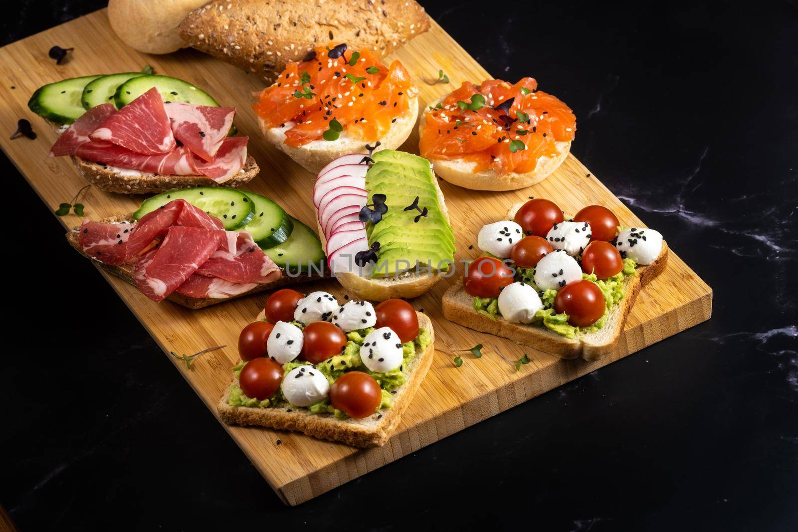 An assortment of sandwiches with fish, cheese, meat and vegetables lay on the board and a bun by Lobachad