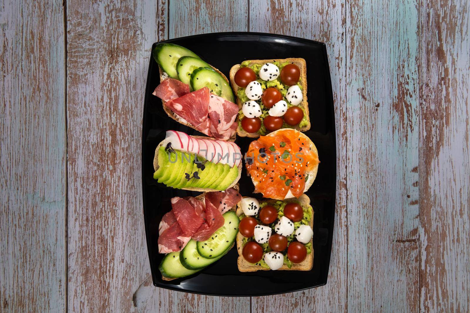 Assorted sandwiches with fish, cheese, meat and vegetables on a black plate and wooden background by Lobachad