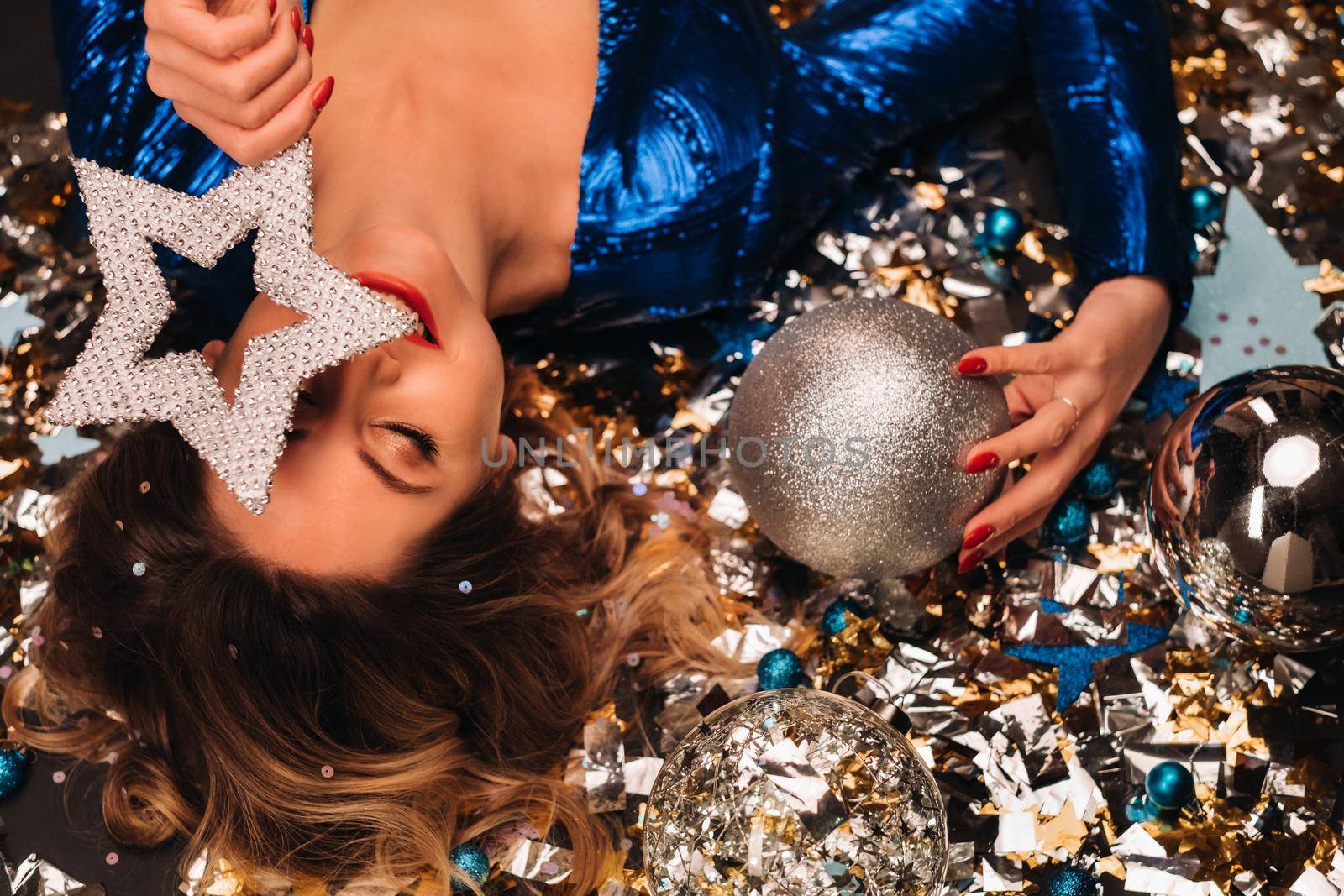 a woman in a blue sequined dress smiles and lies on the floor under a falling multicolored confetti.