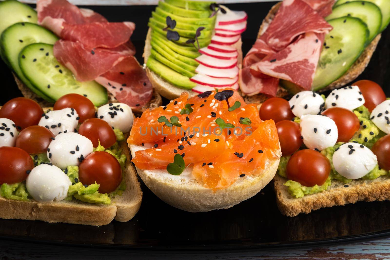 Assorted sandwiches with fish, cheese, meat and vegetables on a black plate and wooden background.