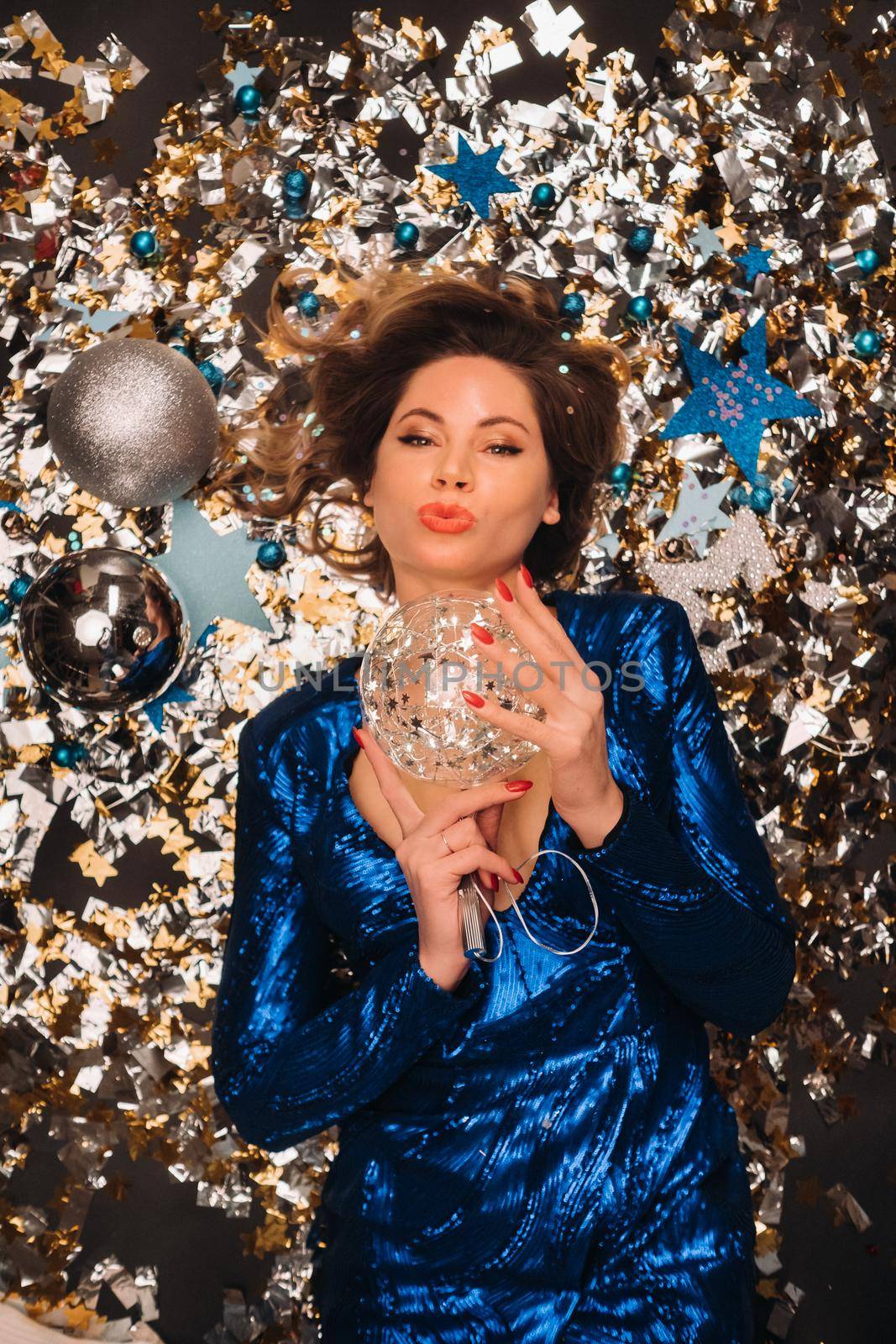 a woman in a blue sequined dress smiles and lies on the floor under a falling multicolored confetti.