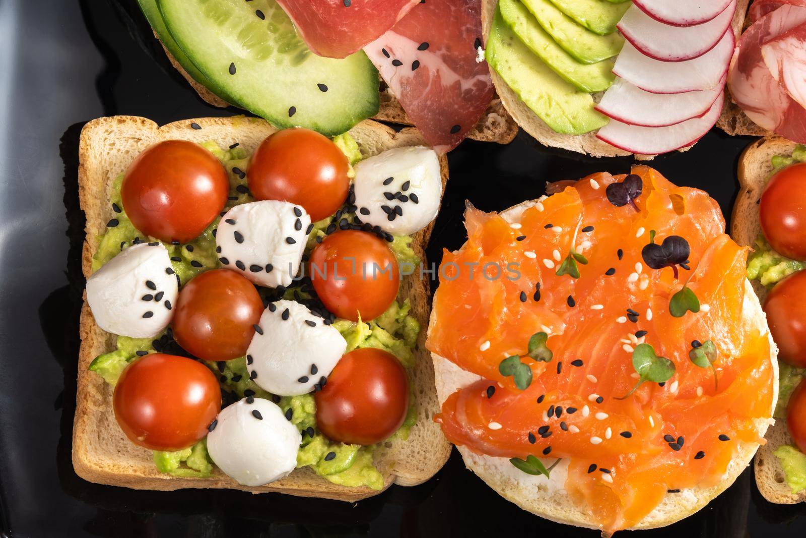 Assorted sandwiches with fish, cheese, meat and vegetables on a black plate.