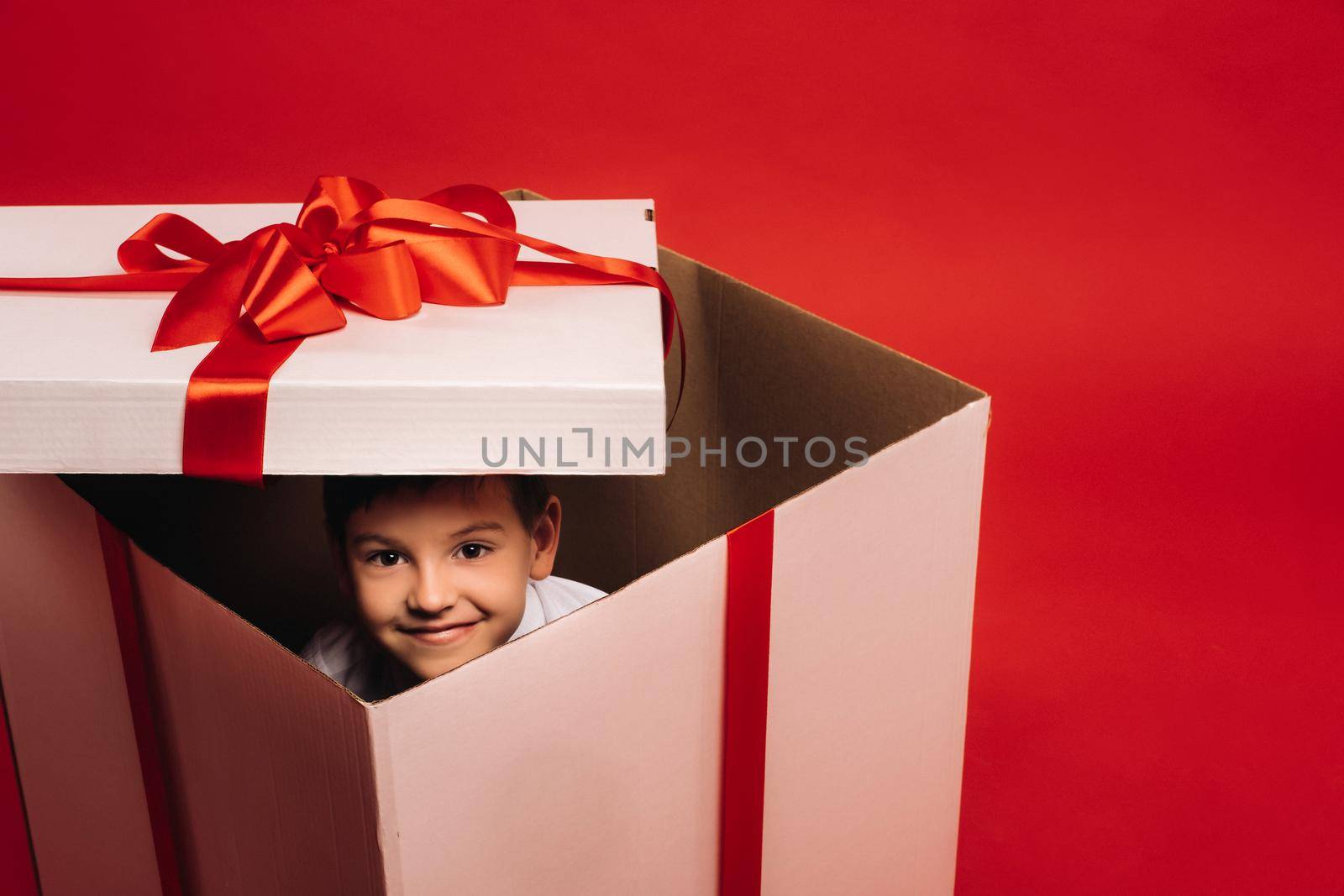 A little boy sits in a Christmas present and looks out of it on a red background.