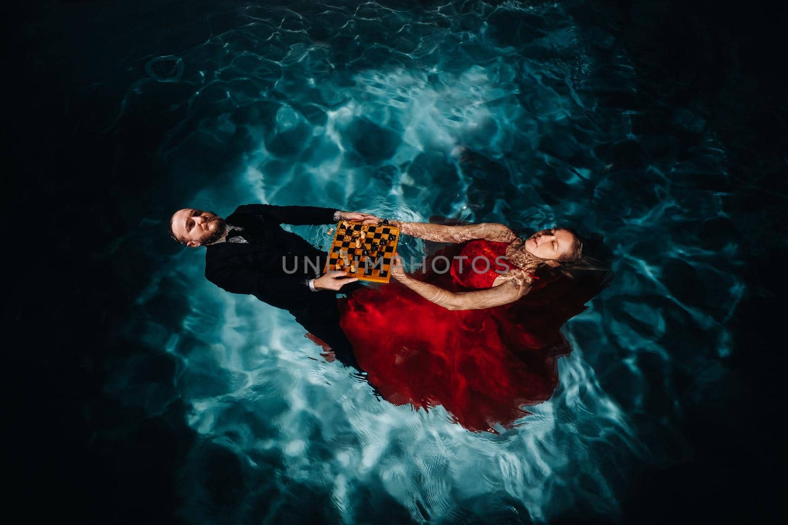 a man in a suit and a girl in a red dress are lying on the water with chess in the pool.