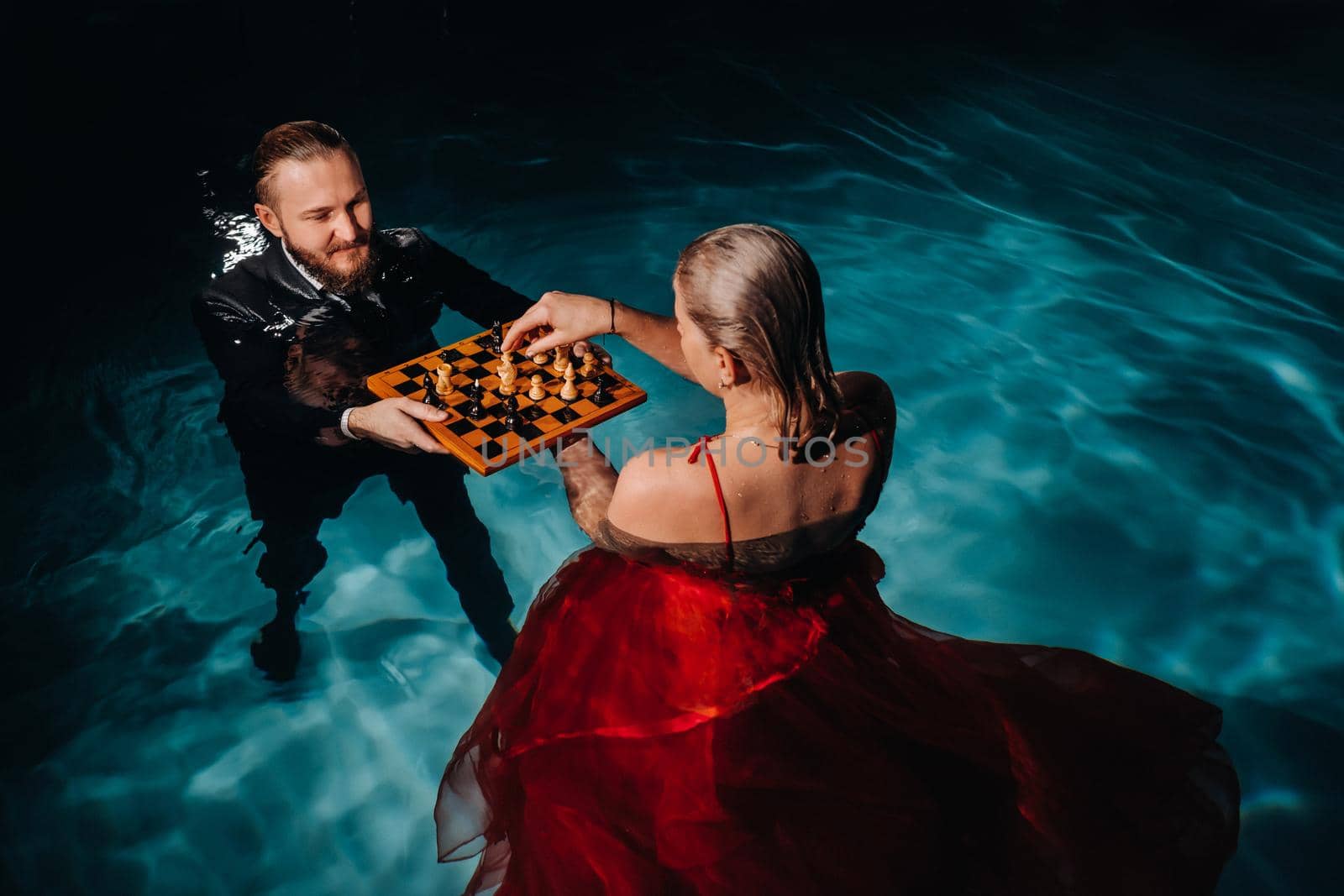 a man in a suit and a girl in a red dress play chess on the water in the pool.