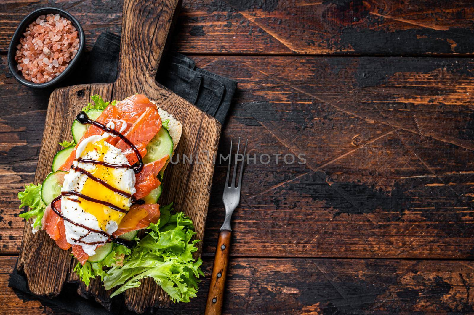 Smoked salmon Sandwich with Benedict egg and avocado on bread. Dark wooden background. Top view. Copy space by Composter