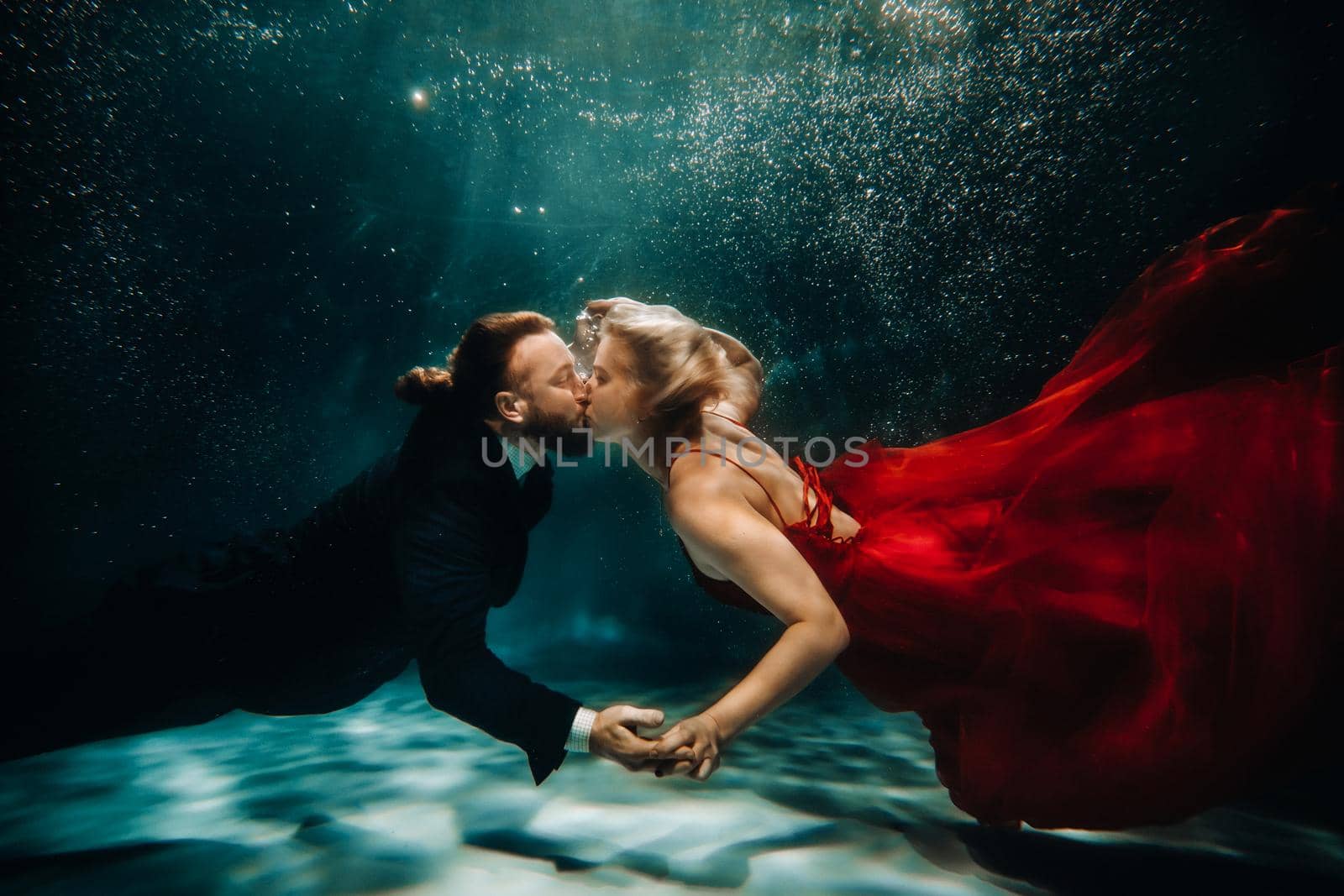 A woman in a red dress and a man in a suit are kissing underwater.A pair of floats under water.