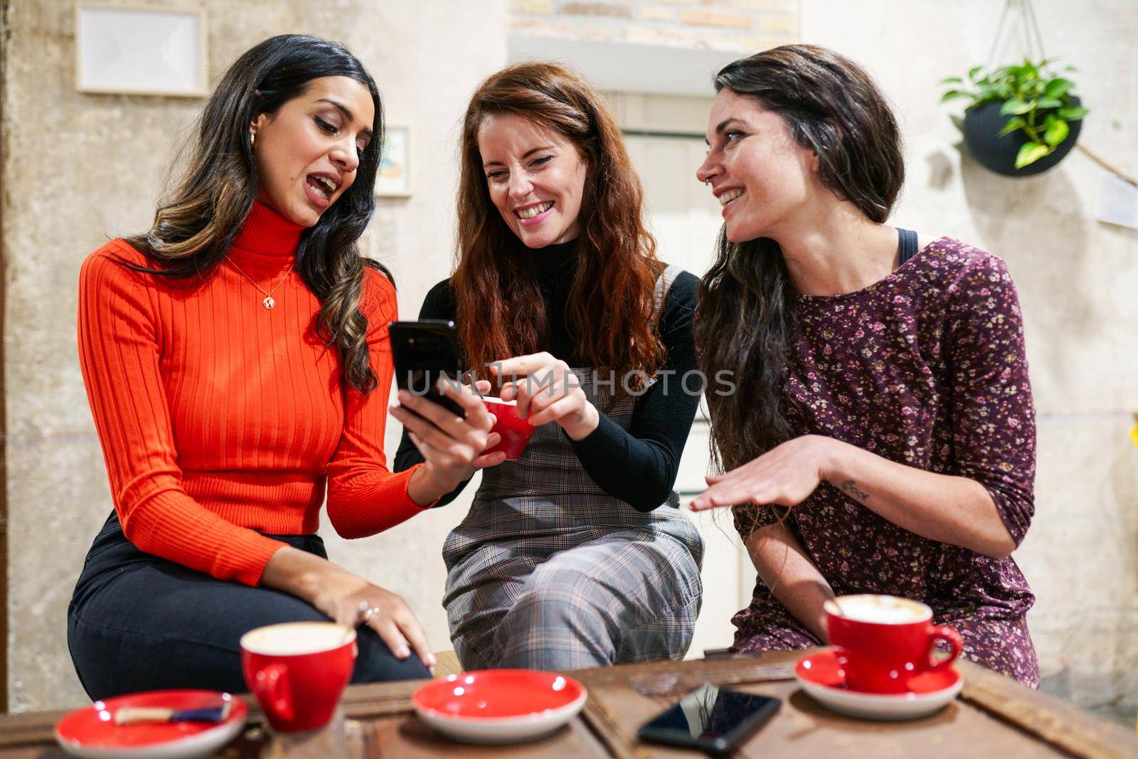 Multiethnic group of three happy female friends looking to a smartphone in a cafe bar.