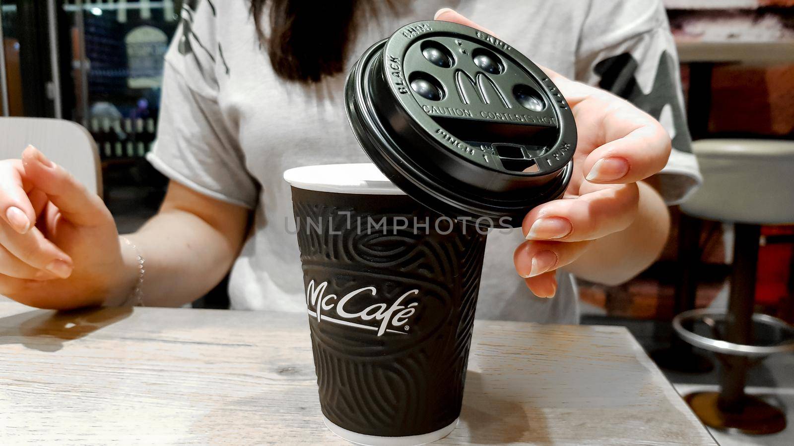 Ukraine, Kiev - September 09, 2019: A girl covers a black plastic paper lid with a hot black coffee maker in McDonalds, McCafe sitting at a table in a fast food restaurant.