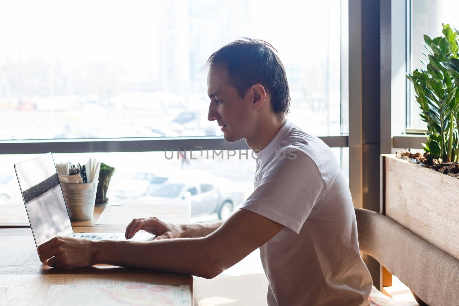 Young businessman holding mug of coffee while working on laptop computer in store with healthy food, male freelancer writing text on laptop keyboard while enjoying cup of cappuccino in modern cafe