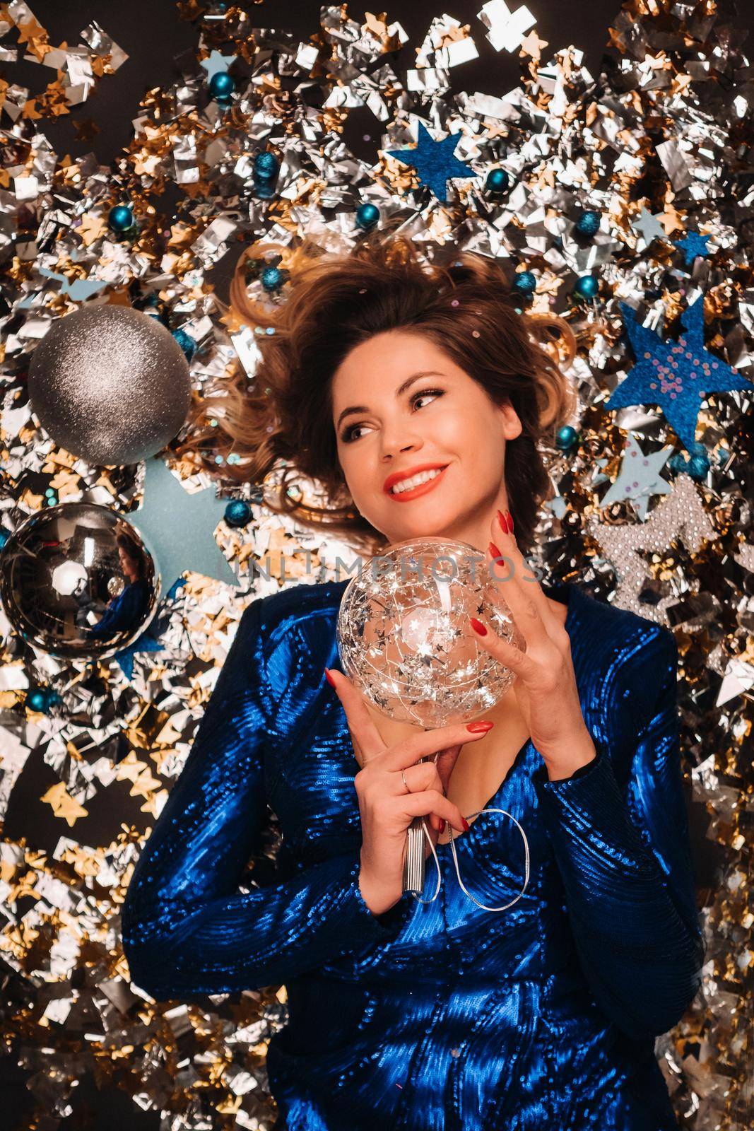 a woman in a blue sequined dress smiles and lies on the floor under a falling multicolored confetti.
