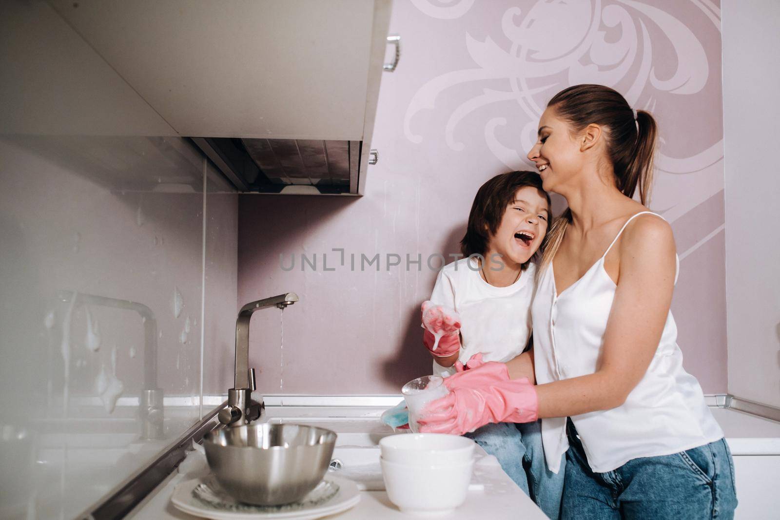 housewife mom in pink gloves washes dishes with her son by hand in the sink with detergent. A girl in white and a child with a cast cleans the house and washes dishes in homemade pink gloves.A child with a cast washes dishes and smiles by Lobachad