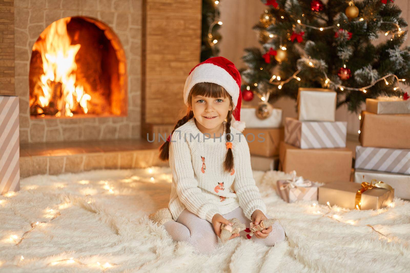 Charming kid in Santa hat and white sweater sitting on carpet near christmas tree and fireplace wit tiny toys in hands, looks smiling at camera, celebrating holiday at home.