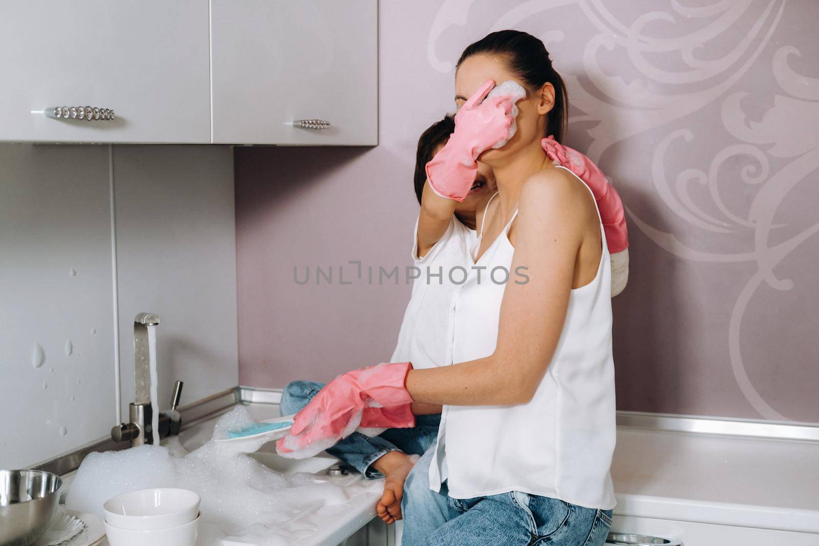 housewife mom in pink gloves washes dishes with her son by hand in the sink with detergent. A girl in white and a child with a cast cleans the house and washes dishes in homemade pink gloves.A child with a cast washes dishes and smiles by Lobachad