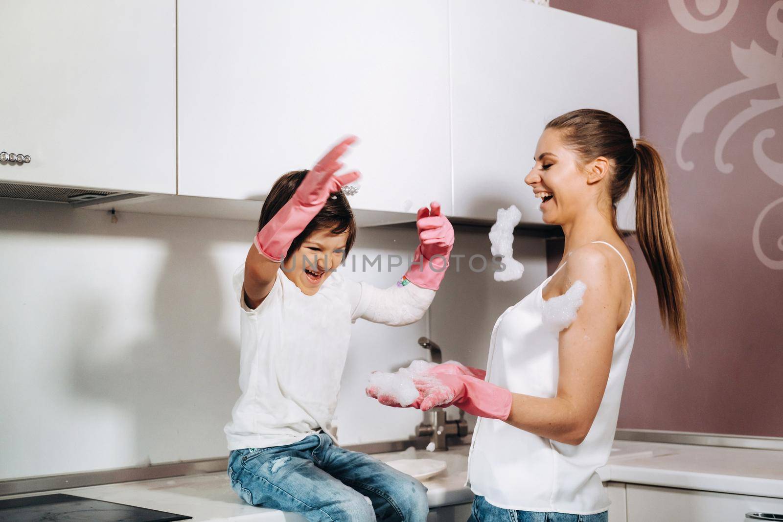 housewife mom in pink gloves washes dishes with her son by hand in the sink with detergent. A girl in white and a child with a cast cleans the house and washes dishes in homemade pink gloves.A child with a cast washes dishes and smiles by Lobachad