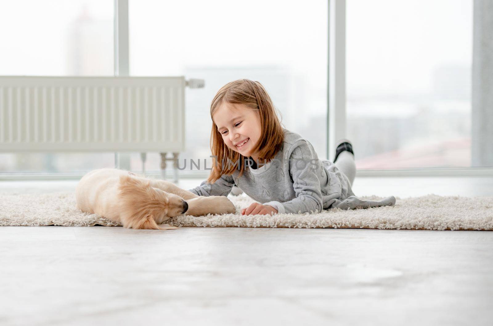 Smiling little girl with young dog golden retriever lying in light room