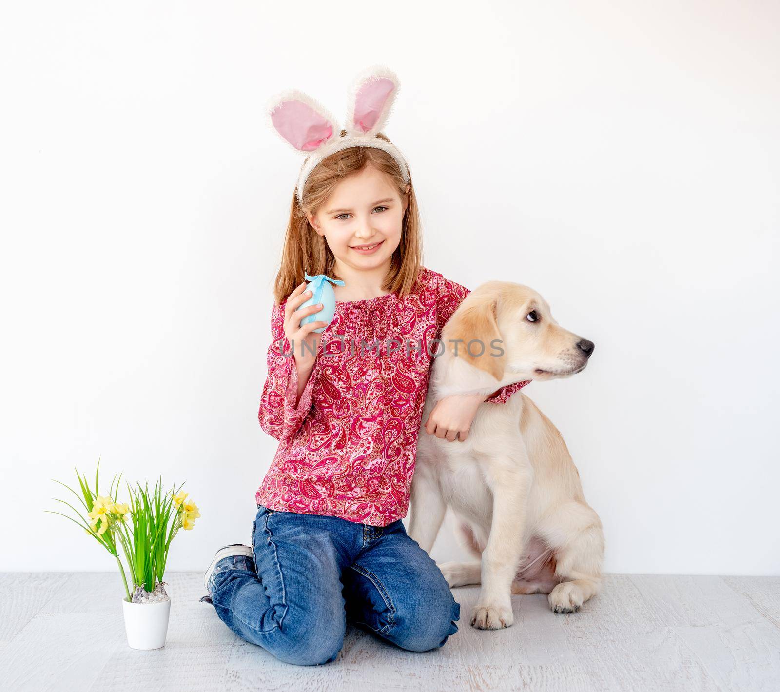 Funny little girl in bunny ears holding colored egg and hugging young dog in light interior