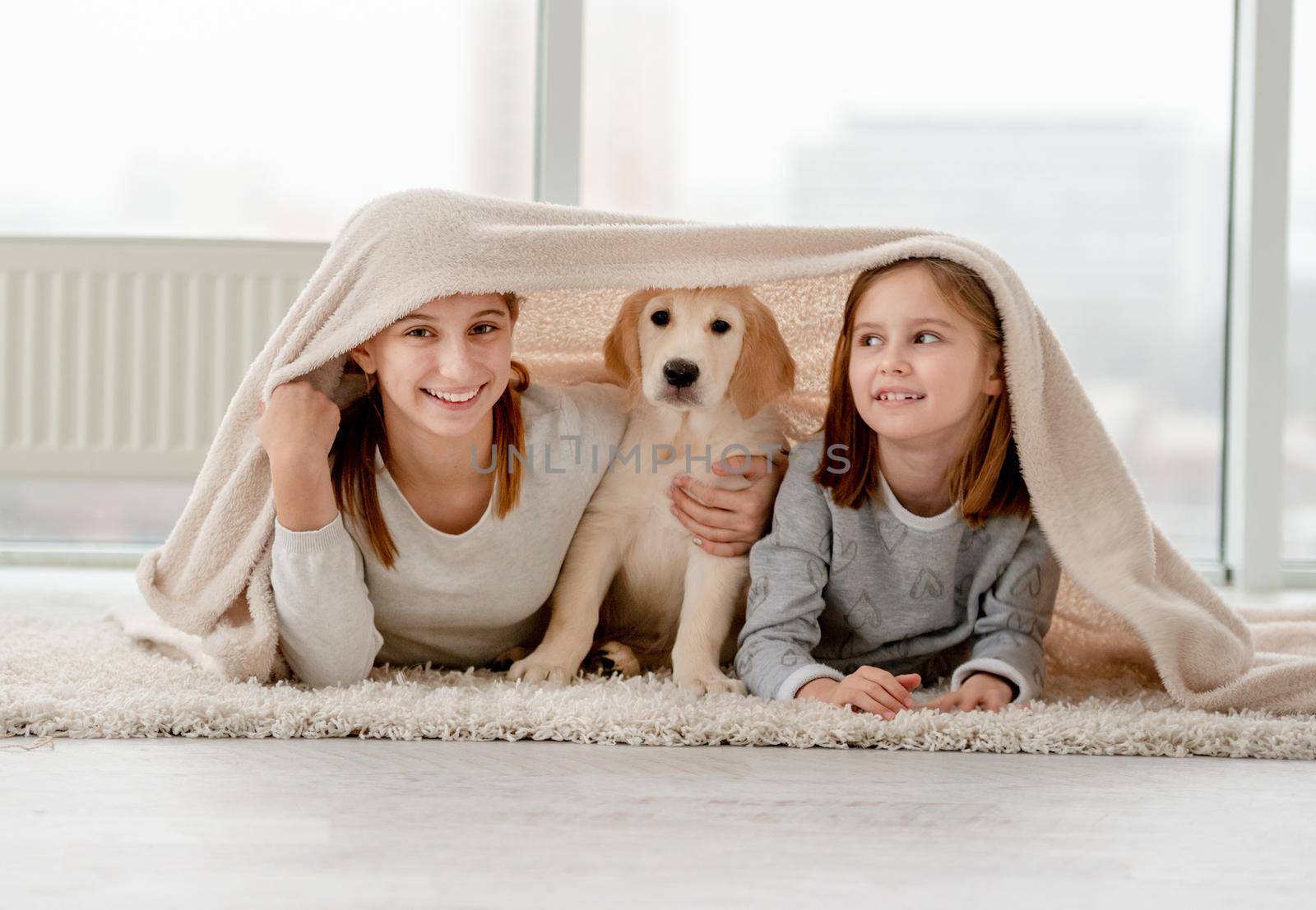 Lovely sisters and golden retriever puppy under plaid on floor in room