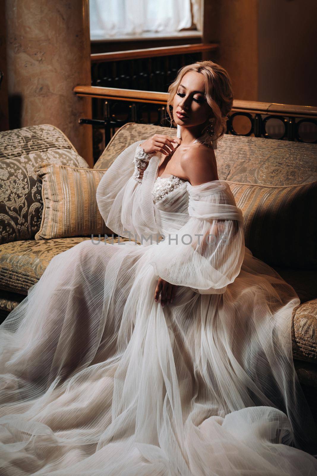 A beautiful bride is sitting on a luxurious sofa. Beautiful model girl in a white wedding dress. Female portrait of a nice lady. Woman with hair.