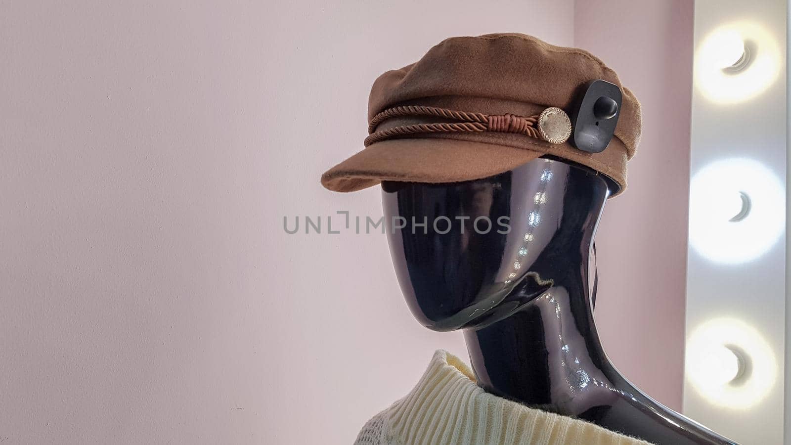 Portrait of a black mannequin with a brown hat in a women fashion store. Mannequin woman in a hat on a pink wall background and a large white mirror with backlight and light bulbs.