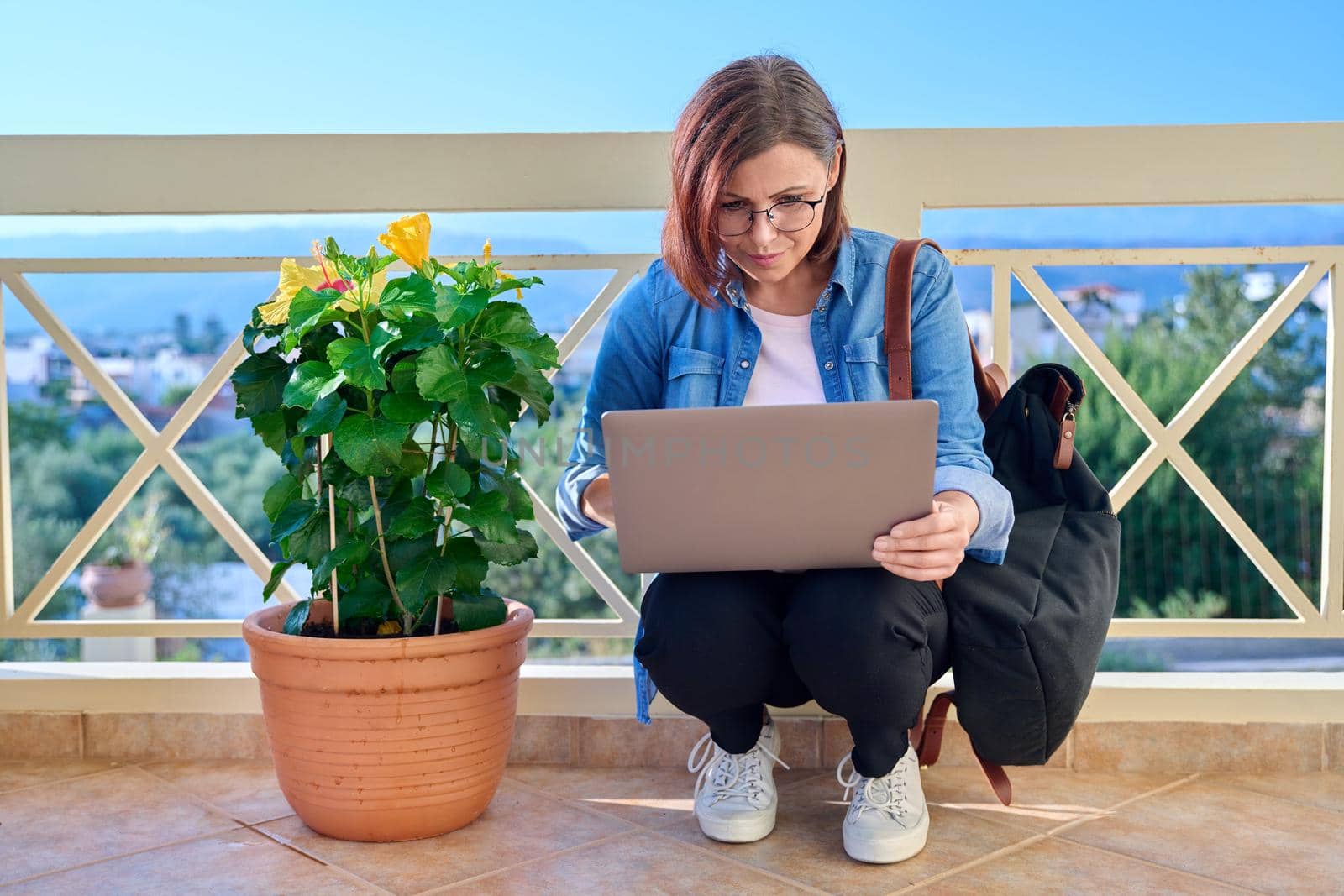 Confident serious mature businesswoman with laptop outdoors. Middle-aged female in denim shirt headphones glasses with backpack looking at laptop screen. Work, business, technology, people 40s age