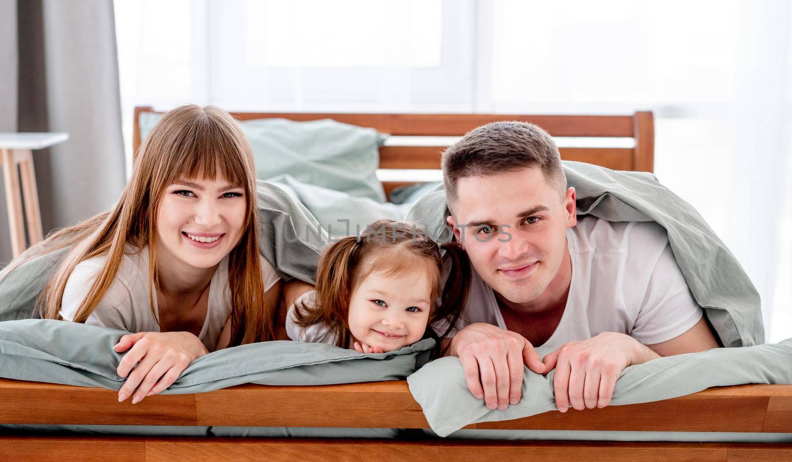 Family under the blanket in the bed. Young parents playing with small daughter under coverlet and smiling. Beautiful girl with her husband and kid in the bedroom