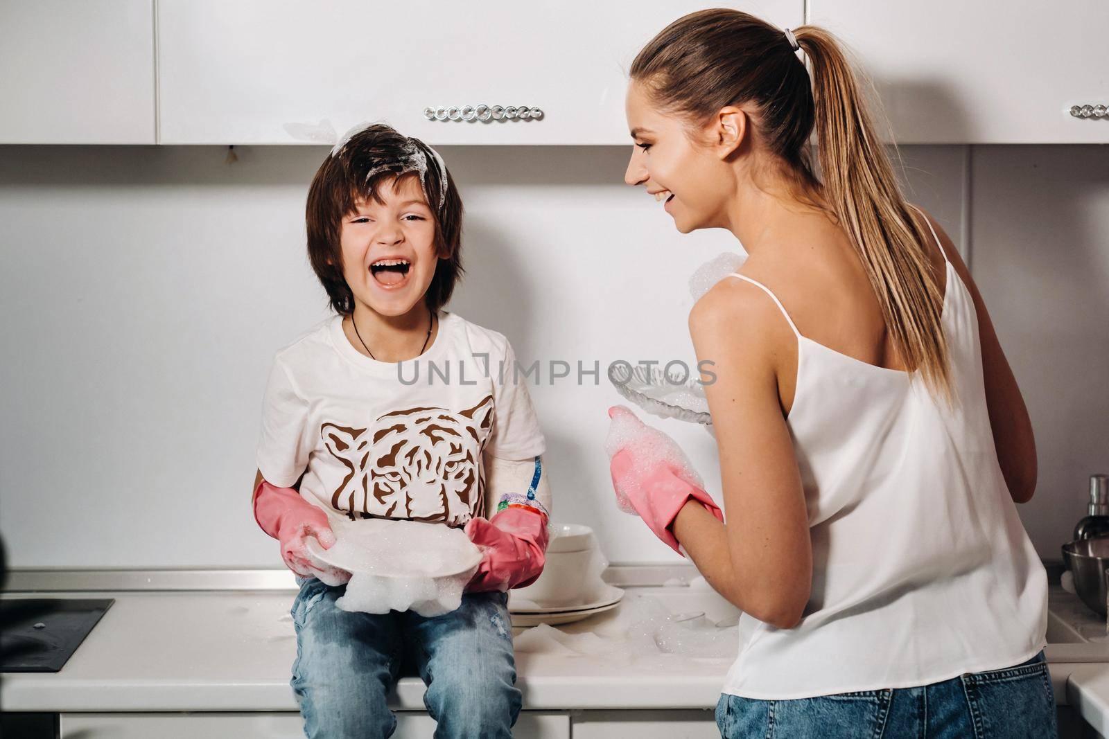 housewife mom in pink gloves washes dishes with her son by hand in the sink with detergent. A girl in white and a child with a cast cleans the house and washes dishes in homemade pink gloves.A child with a cast washes dishes and smiles.