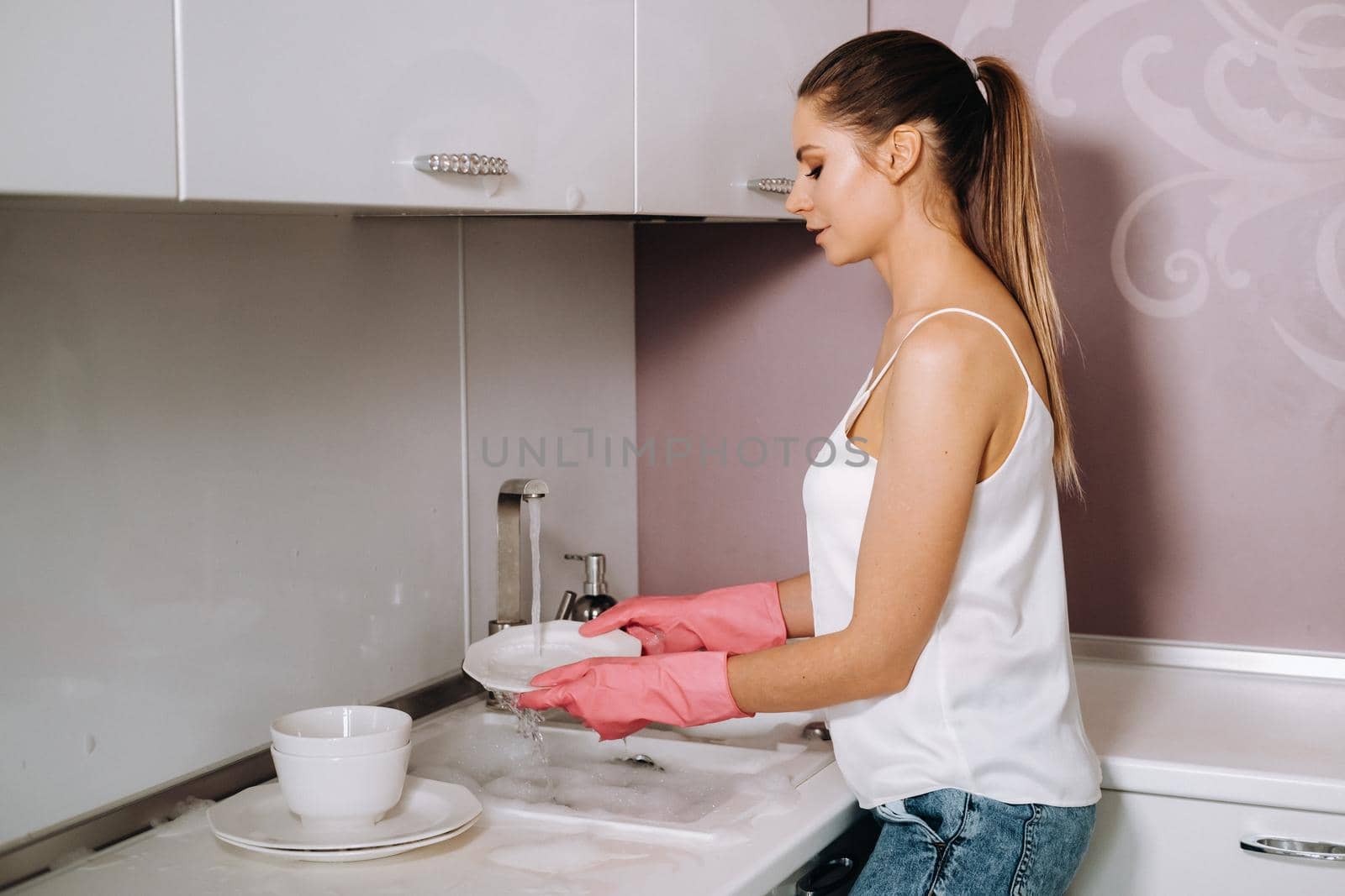 housewife girl in pink gloves washes dishes by hand in the sink with detergent. The girl cleans the house and washes dishes in gloves at home. by Lobachad