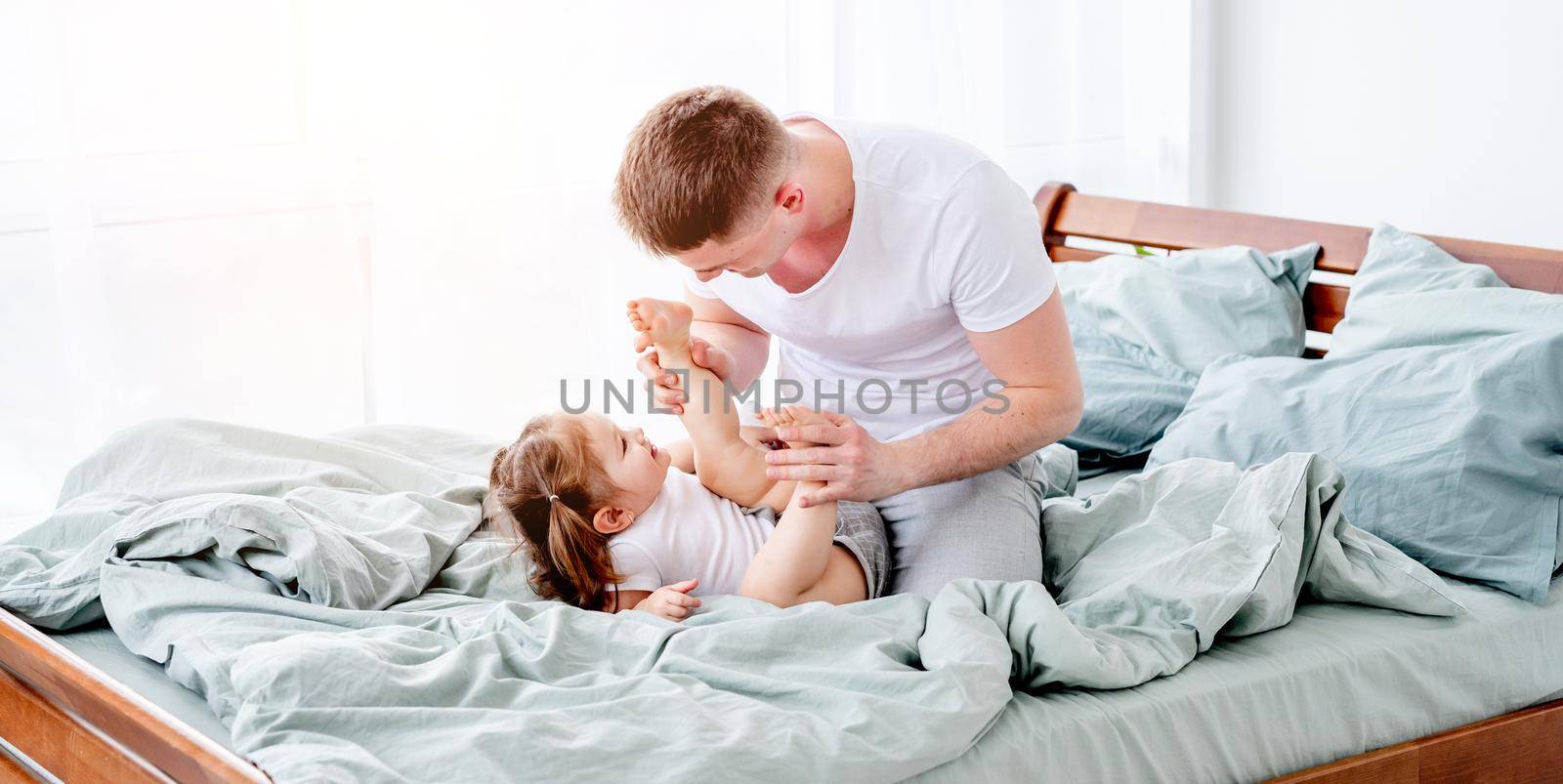 Young father spending morning with his little daughter lying in the bed. Smiling child with her parent in sunny room. Beautiful moments of dad with his kid in the bedroom together