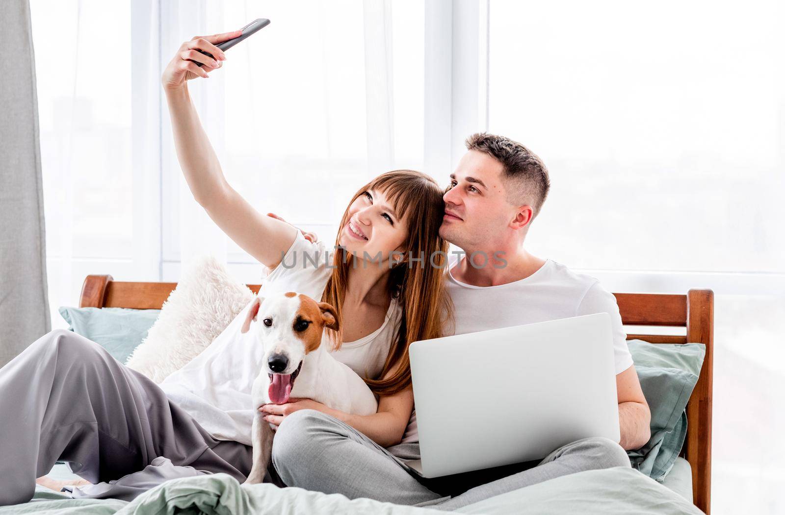 Young couple with cute dog in the bed making selfie. Beautiful woman and man with laptop do photos using smartphone. Family morning with pet and technologies