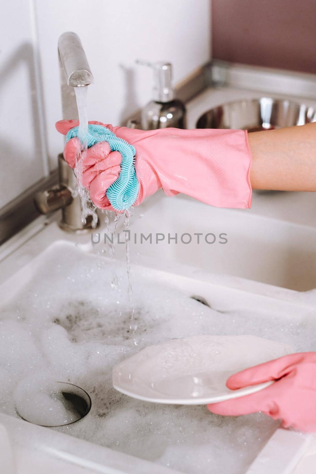 housewife girl in pink gloves washes dishes by hand in the sink with detergent. The girl cleans the house and washes dishes in gloves at home. by Lobachad