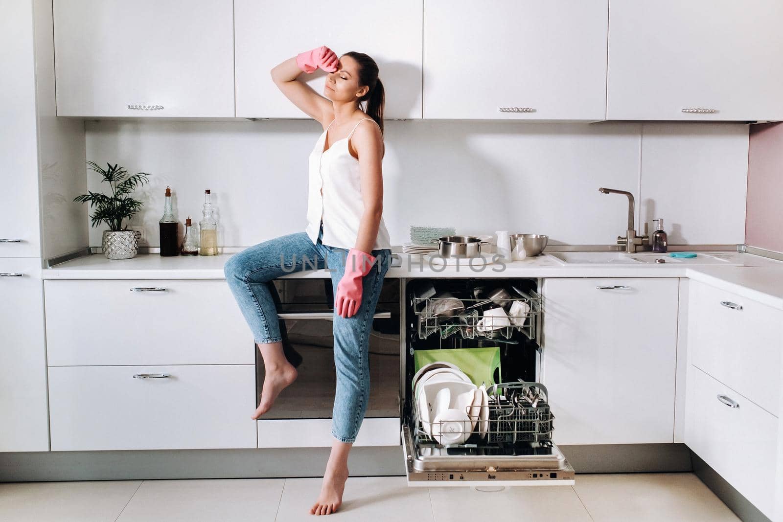 a housewife girl in pink gloves after cleaning the house sits tired in the kitchen.In the white kitchen, the girl has washed the dishes and is resting.Lots of washed dishes.