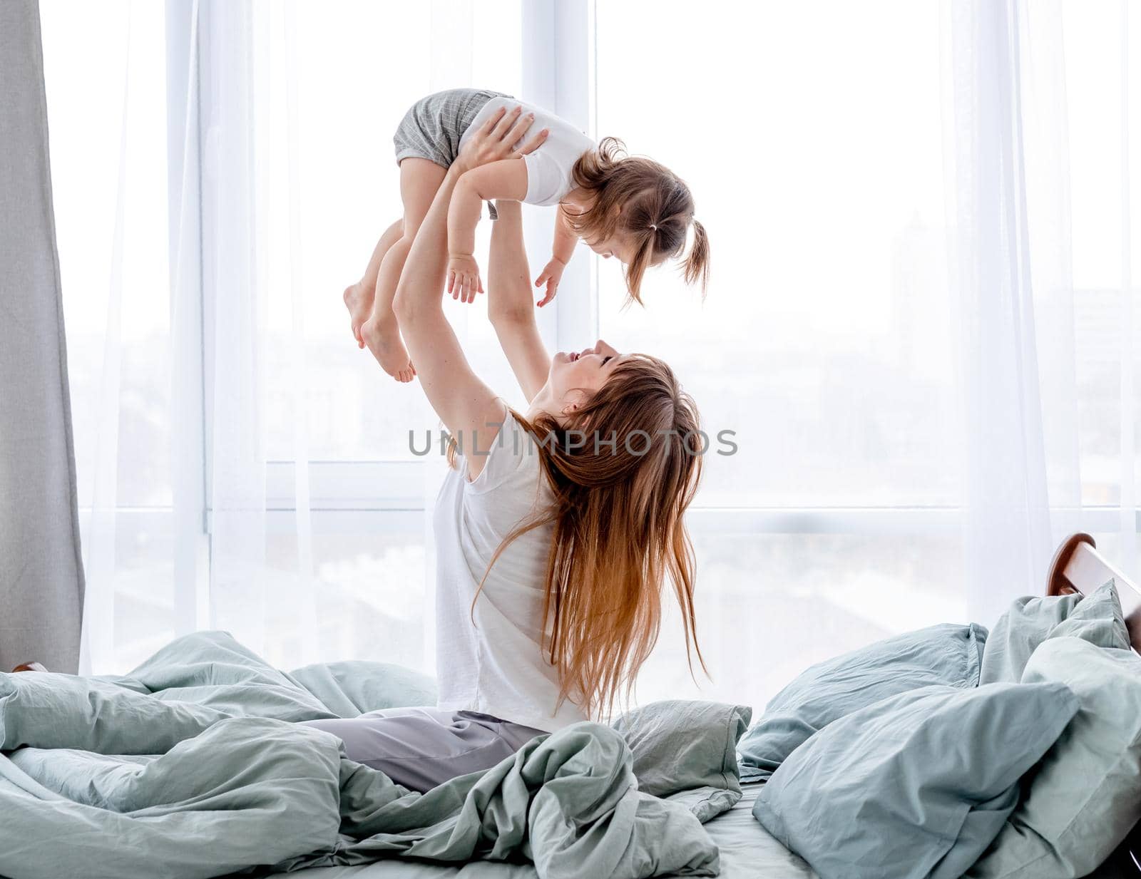 Young mother playing with little cute daughetr in the bed holding her in hands. Beautiful family morning together. Lovely moments of mom with her child