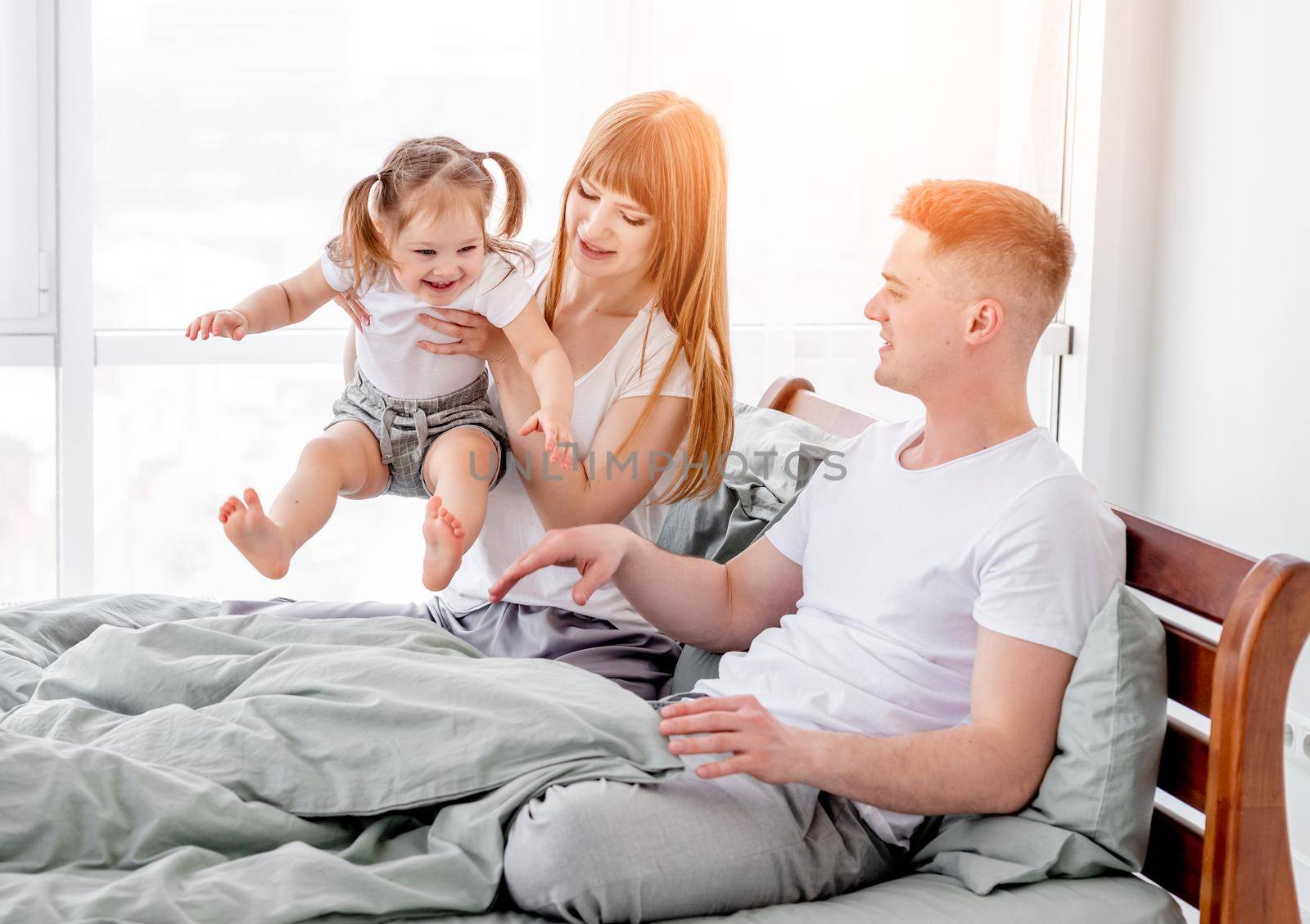 Sunny family mornents in the bedroom. Beautiful parents with their daughter staying in the bed and enjoying time together. Mother holding her smiling child in her hands