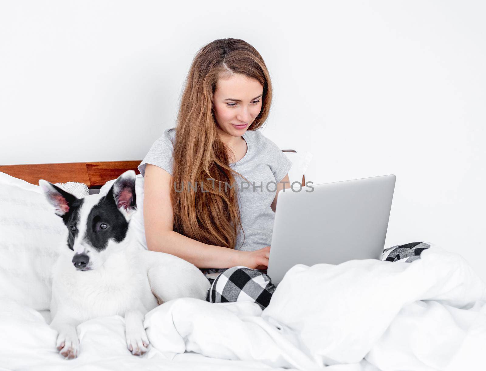 Beautiful girl wearing pajamas sitting in the bed with laptop working and cute doggy lying close to her. Young woman freelancer at home with pet in morning time typing on notebook. Remote work concept