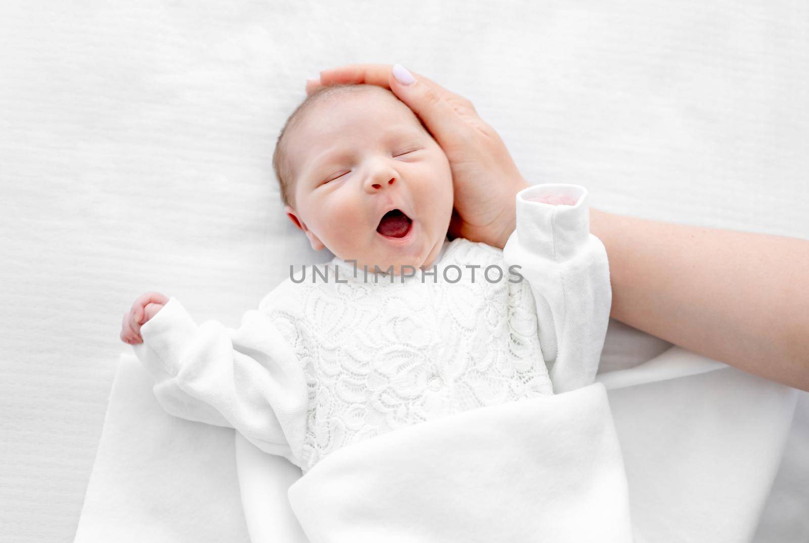 Adorable newborn baby girl wearing white costume lying in the bed at home with daylight, yawning during sleeping and her mother hand cares about her. Portrait of cute infant child and parent love