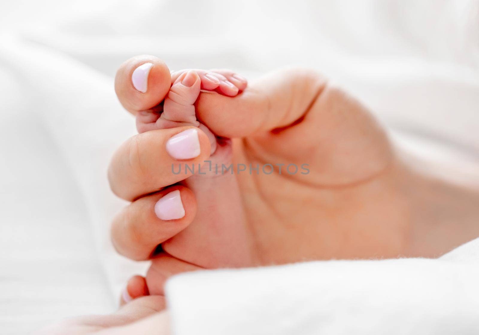 Mother holding tiny newborn baby hand with little fingers closeup in the bed with white bedding. Concept of matherinity love, care and tenderness