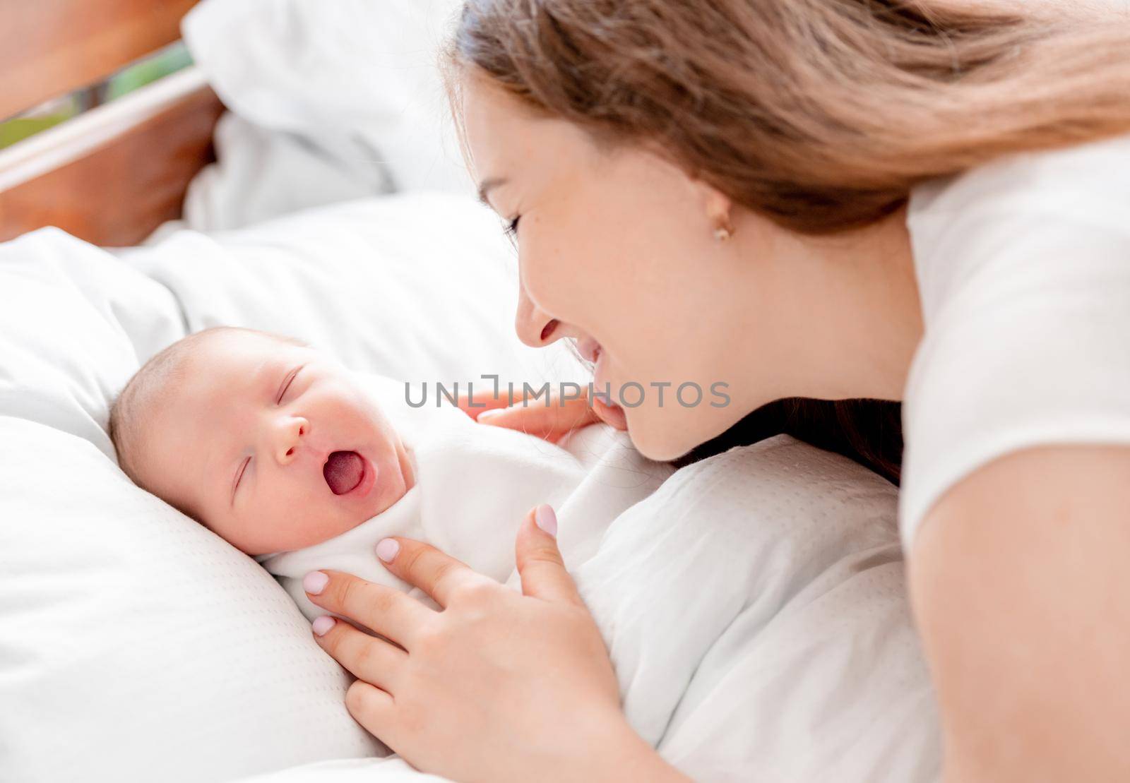 Young mother looking at her sleeping with opened mouth newborn daughter swaddled in white sheets at home and smiling with tenderness. Portrait of girl mom with her infant child napping in the bed