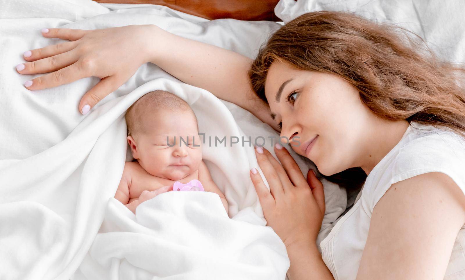Beautiful young mother lying in the bed with newborn daughter and looking at her. Adorable infant baby girl sleeping close to her mom parent