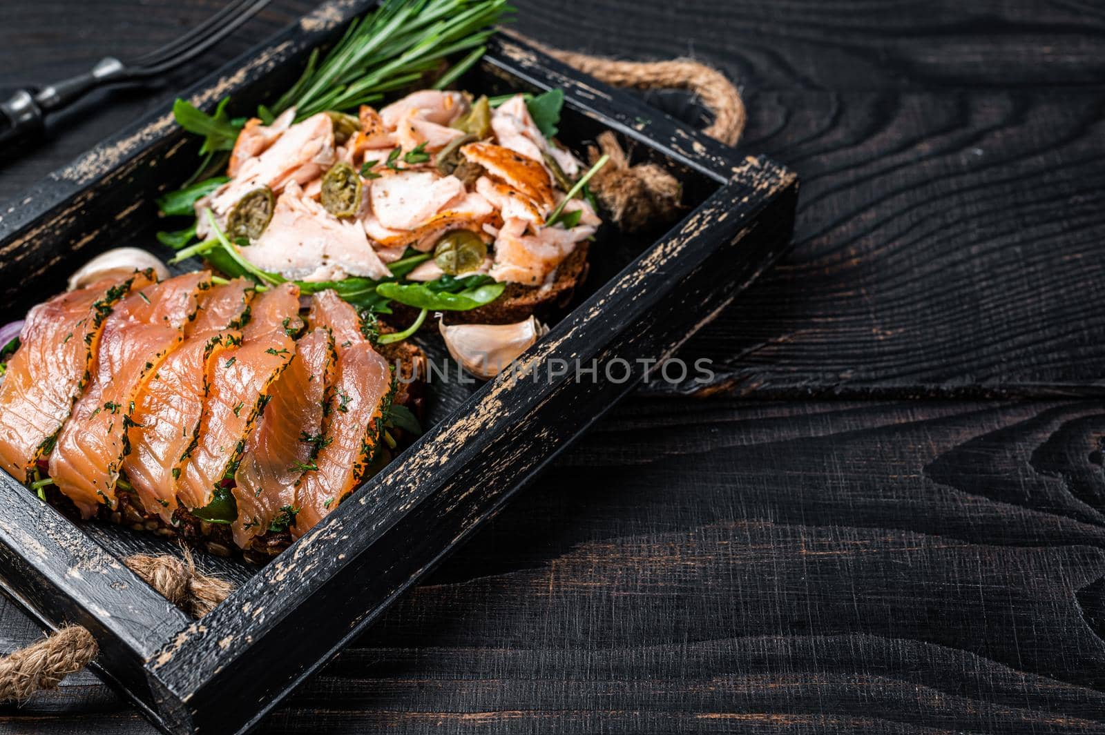 Toasts with hot and cold smoked salmon, arugula in a wooden tray with herbs. Black wooden background. Top view. Copy space by Composter