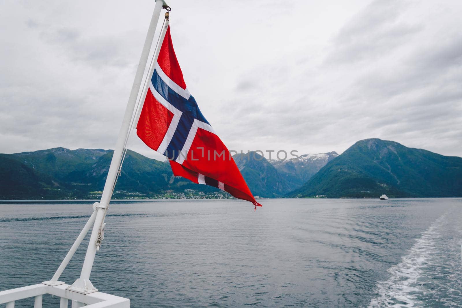 Norwegian flag hanging on the railing of the ship and waving above the water. Norvegian fjord with a flag. Ferry trip in Norway. Norway Flag on sea and mountains background by Tomashevska