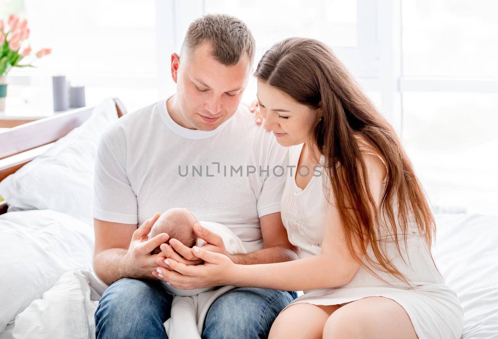 Beautiful young family sitting in the bed and holding newborn daughter in the room with daylight. Adorable infant baby girl sleeping in hands of her mother and father at home
