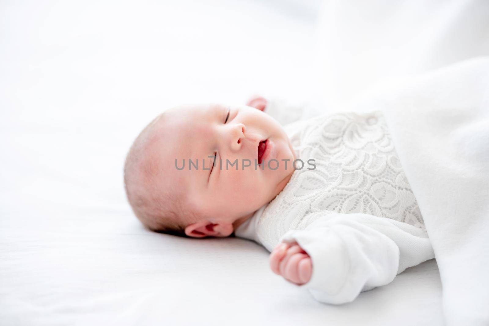 Adorable newborn baby girl wearing white costume lying in the bed and sleeping with mouth open. Cute infant child napping at home
