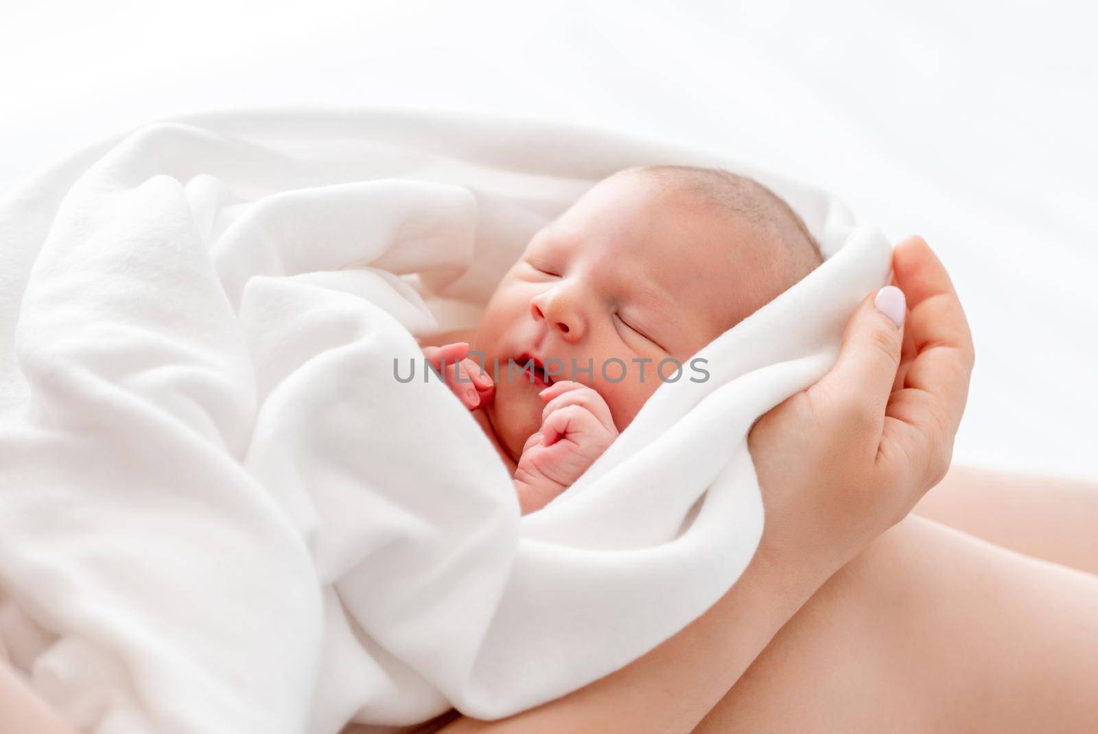 Newborn baby girl swaddled in white sheets sleeping in her mother hands. Closeup portrait of napping infant child with parent at home