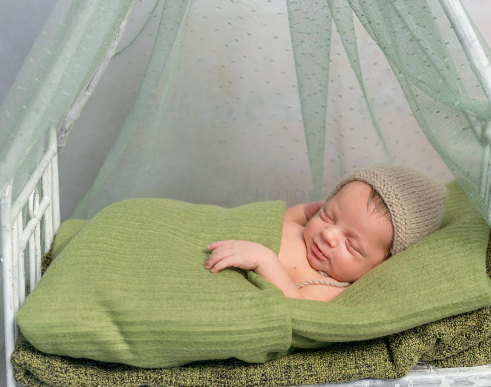 newborn baby in hat covered with green blanket sleeping on little bed with canopy