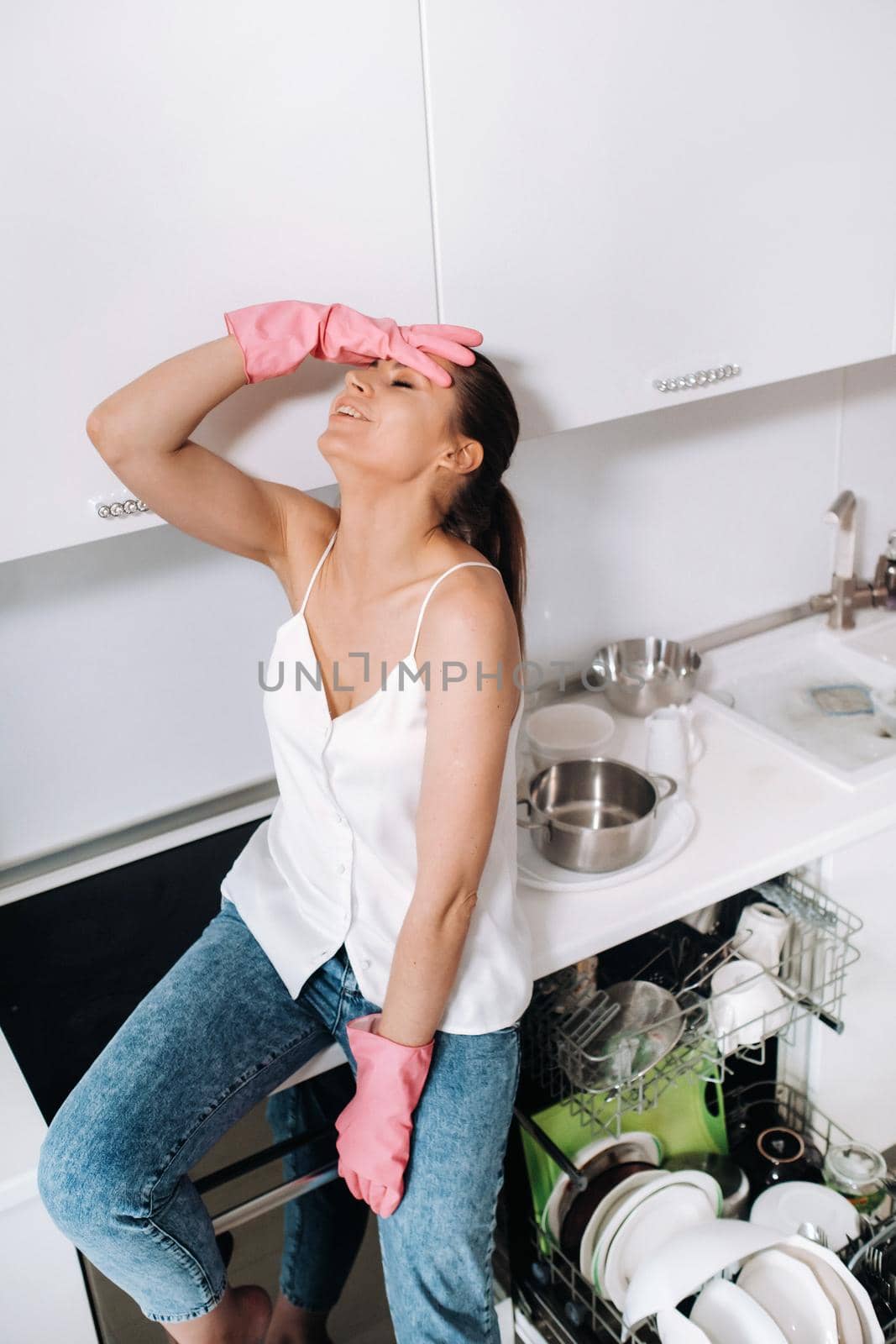 a housewife girl in pink gloves after cleaning the house sits tired in the kitchen.In the white kitchen, the girl has washed the dishes and is resting.Lots of washed dishes.