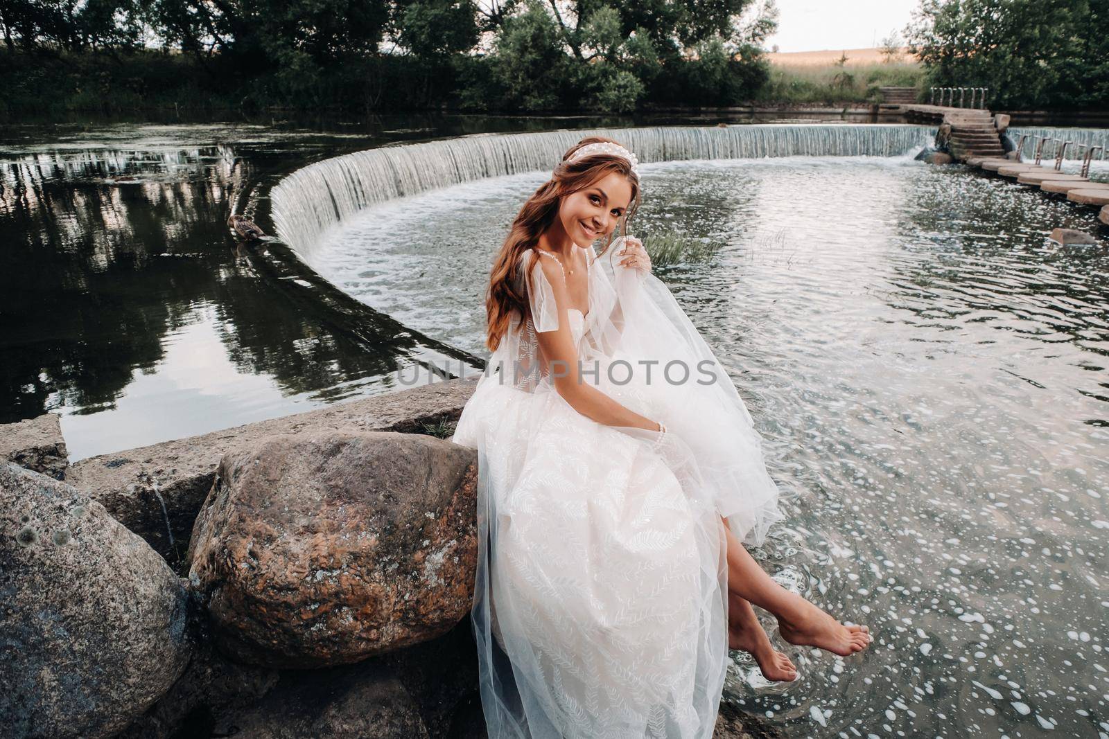 An elegant bride in a white dress, gloves and bare feet is sitting near a waterfall in the Park enjoying nature.A model in a wedding dress and gloves at a nature Park.Belarus.